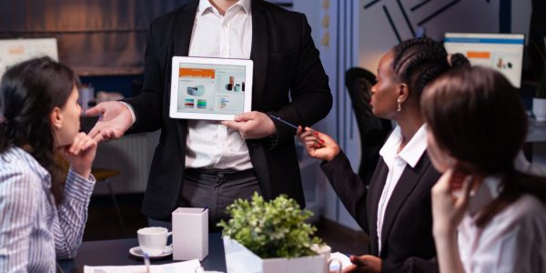 Business entrepreneur man presenting company statistics using tablet for financial presentation. Diverse multi-ethnic businesspeople brainstorming strategy working in office meeting room late at night